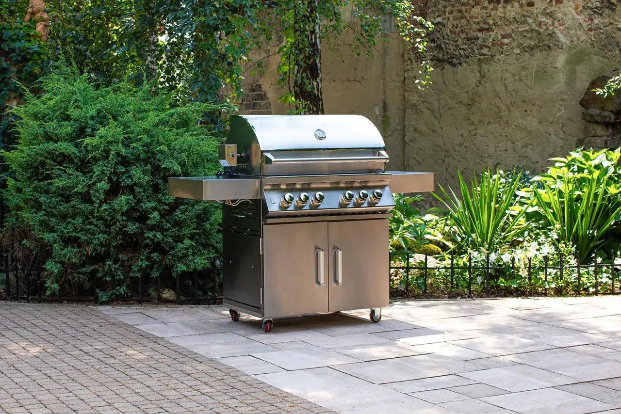 Nowoczesny grill ze stali nierdzewnej stoi na patio, zachowując bezpieczną odległość grilla od budynku. Otoczony zielenią i murem.