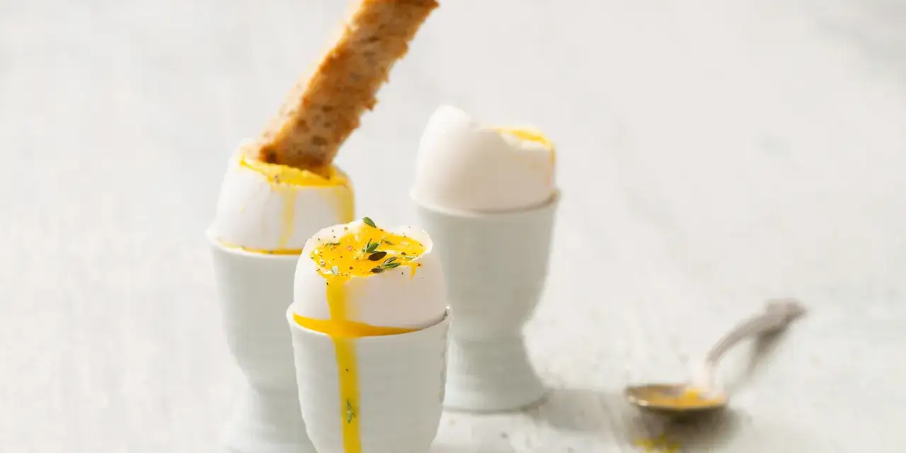 Trois &oelig;ufs &agrave; la coque, dont un avec une mouillette de pain grill&eacute;, sont pr&eacute;sent&eacute;s dans des coquetiers. Le jaune coule d&eacute;licatement.