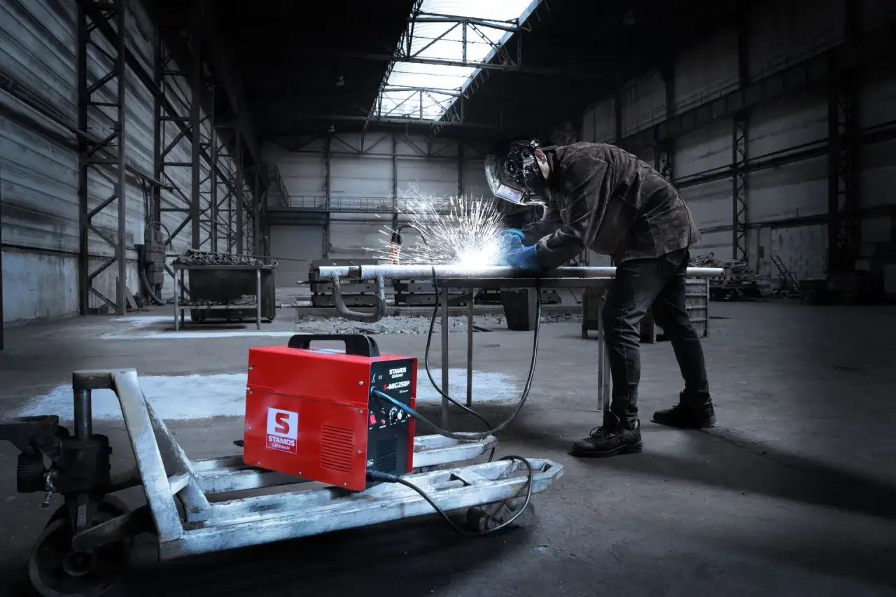 Saldatore al lavoro in un capannone industriale, con una saldatrice rossa STAMOS in primo piano. Utile per capire come scegliere una saldatrice.