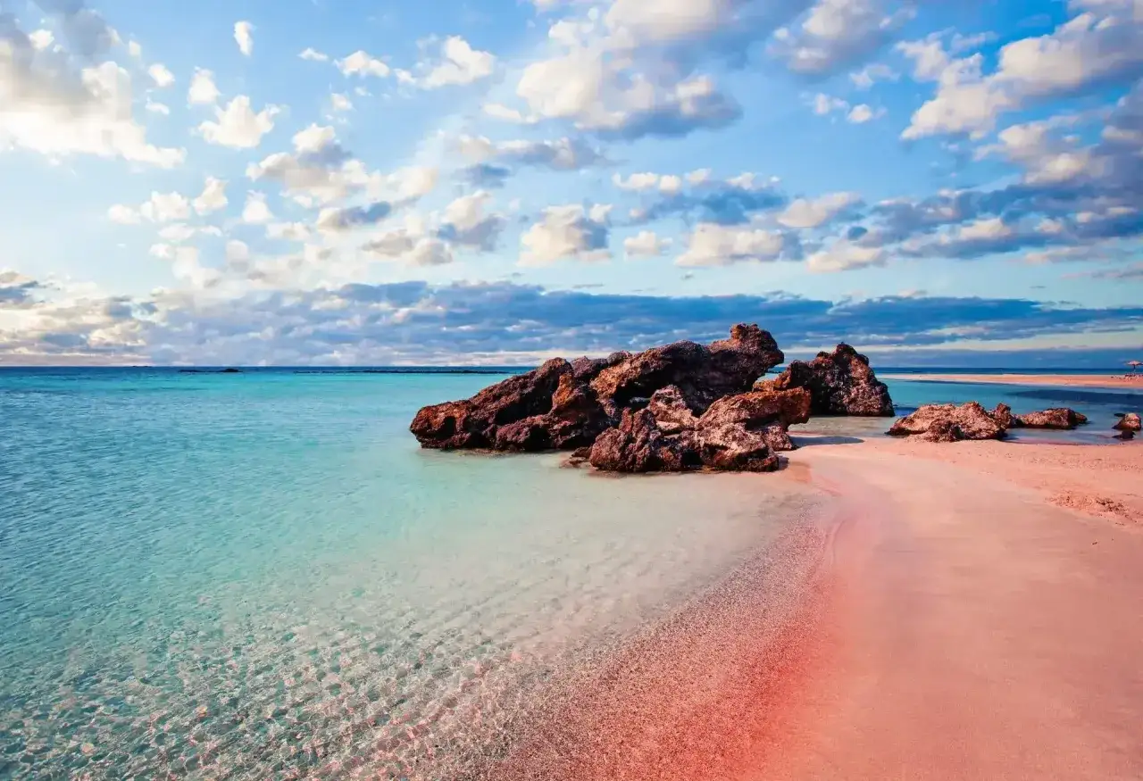 Plage rose de l'ile Crete, eaux turquoise et rochers volcaniques sous un ciel bleu et nuageux.