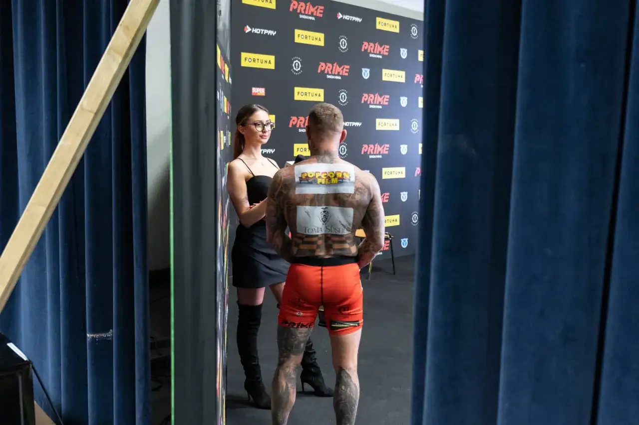 Dziennikarka przeprowadza wywiad z zawodnikiem freak fight, którego plecy zdobią tatuaże i logotypy sponsorów.