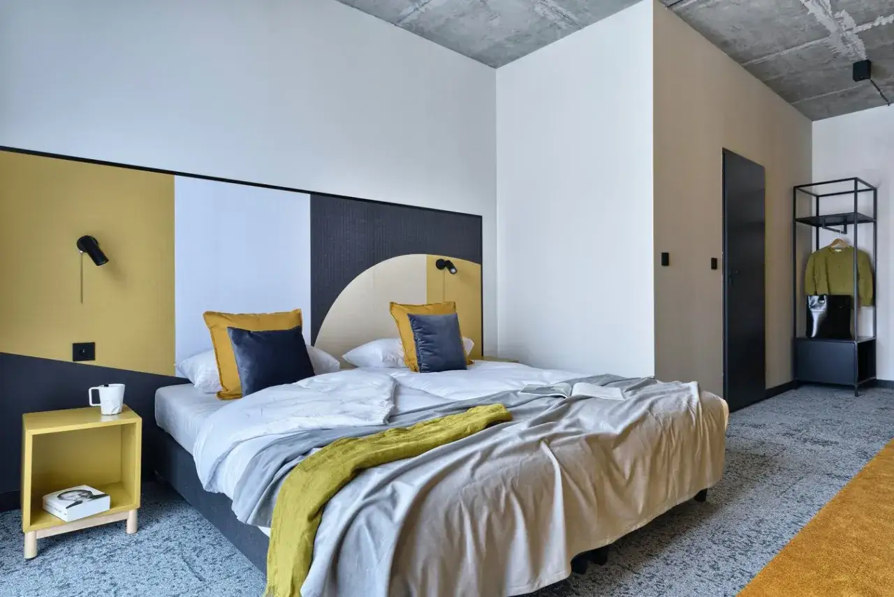 Nowoczesny pok&oacute;j w arche hotel piła. Dwa ł&oacute;żka z ż&oacute;łtymi i granatowymi poduszkami, ż&oacute;łty stolik nocny, czarne drzwi i metalowa szafa.