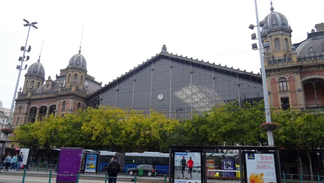 Budapest train station mit seiner beeindruckenden Glasfassade und den historischen Türmen. Bäume säumen den Platz vor dem Bahnhof.