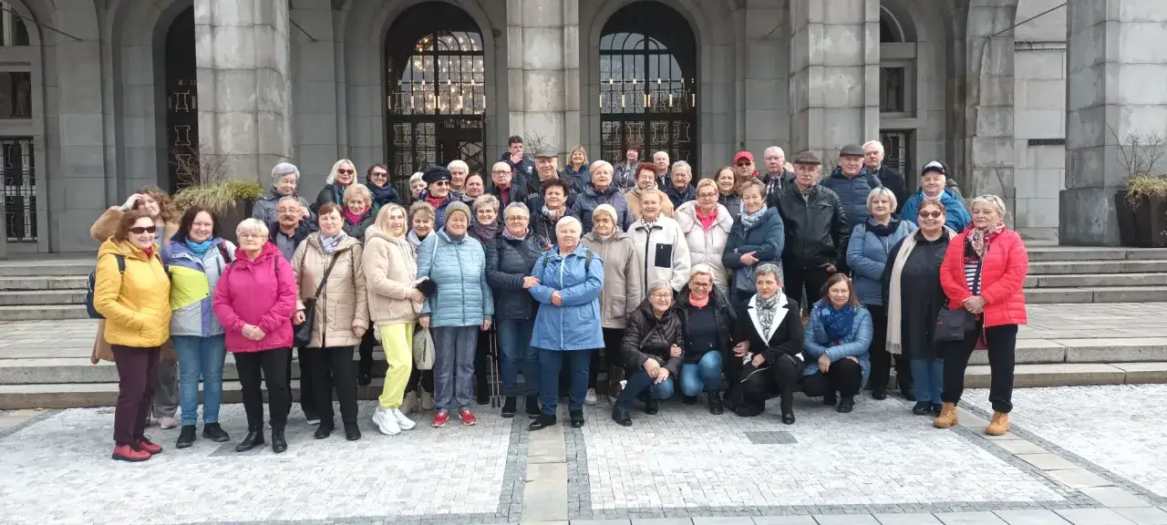 Grupa seniorów pozuje do zdjęcia przed budynkiem. To idealny moment na tanie wycieczki dla seniorów, pełne radości i wspólnych wspomnień.