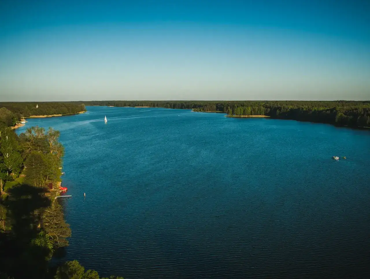 Jezioro na Kaszubach, z czego słyną te tereny – z pięknych krajobrazów i spokoju. Biały żagiel na wodzie, otoczonej lasem.