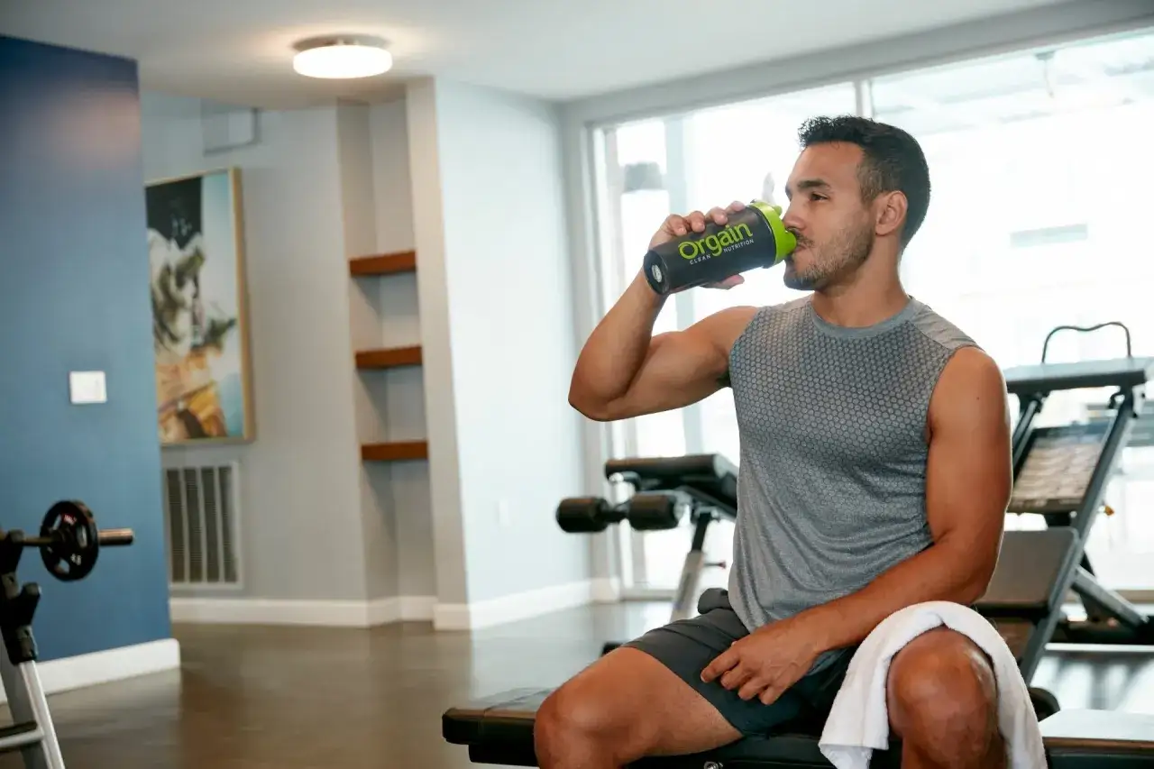 Hombre en el gimnasio bebe prote&iacute;na de Orgain. Un momento ideal para saber **cuando tomar prote&iacute;nas** y optimizar tu entrenamiento.