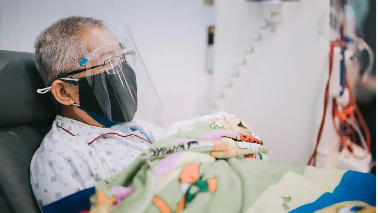 Starszy mężczyzna w masce i przyłbicy, okryty kolorowym kocem, otrzymuje leczenie. Może to być terapia remdesivir.