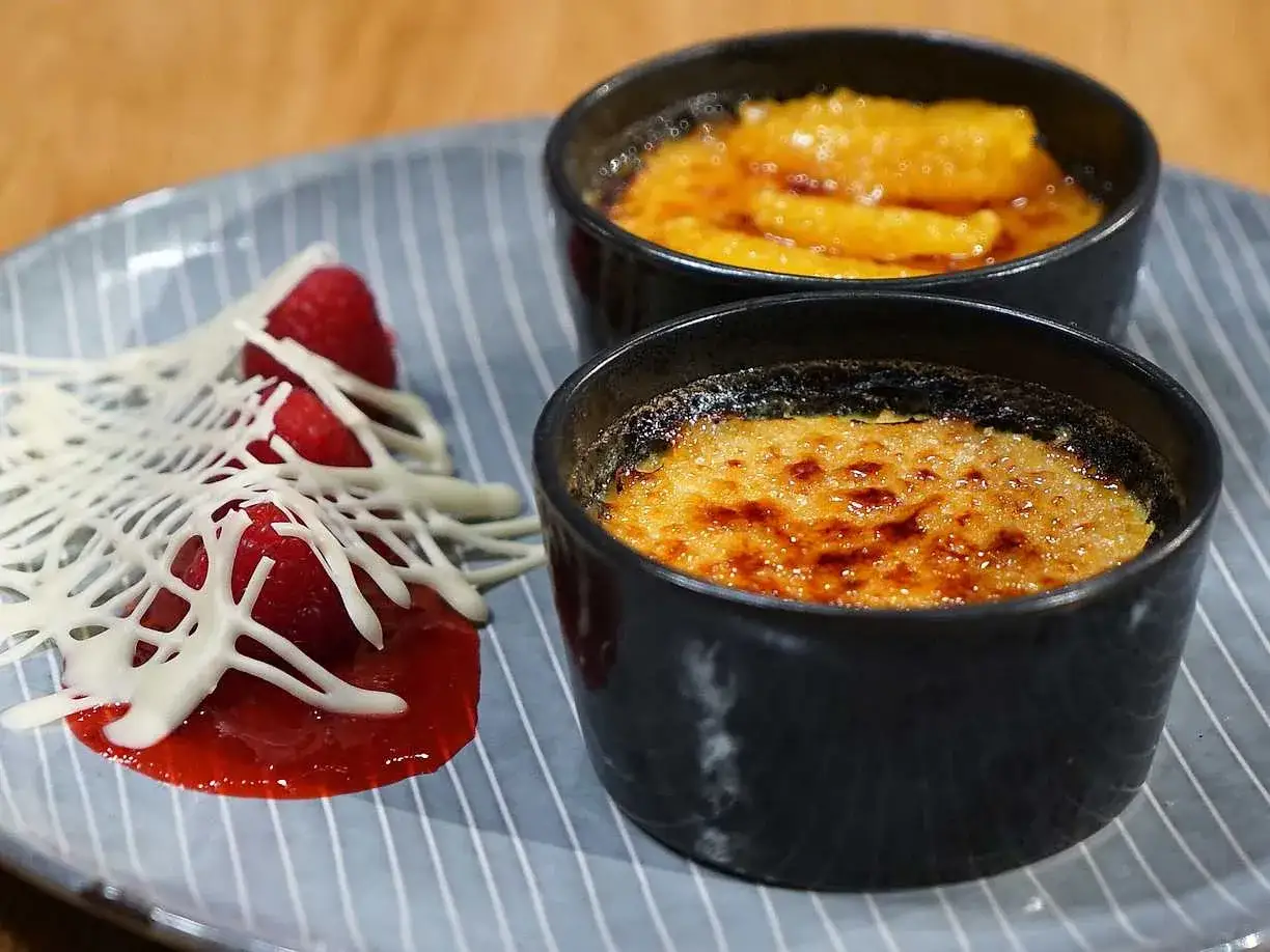 Två krämiga creme brulee med karamelliserad yta, serverade med hallon och vit choklad.