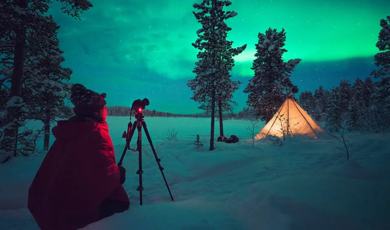 Person i röd jacka förbereder sig att fota norrsken i snöig skog. Ett upplyst tält syns i bakgrunden.