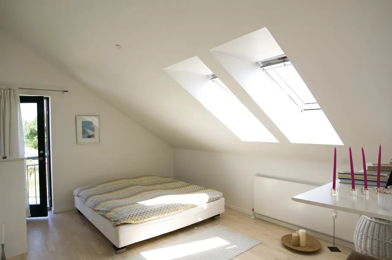 Arredare mansarda: letto accogliente sotto lucernari, porta-finestra con vista sul verde e un tavolino con candele rosa.