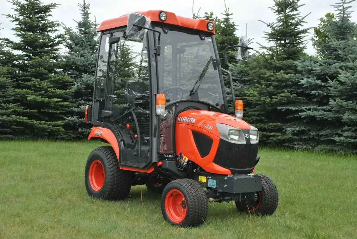 Pomarańczowy traktorek ogrodowy wielofunkcyjny Kubota L231 z kabiną stoi na trawniku, otoczony przez drzewa iglaste.