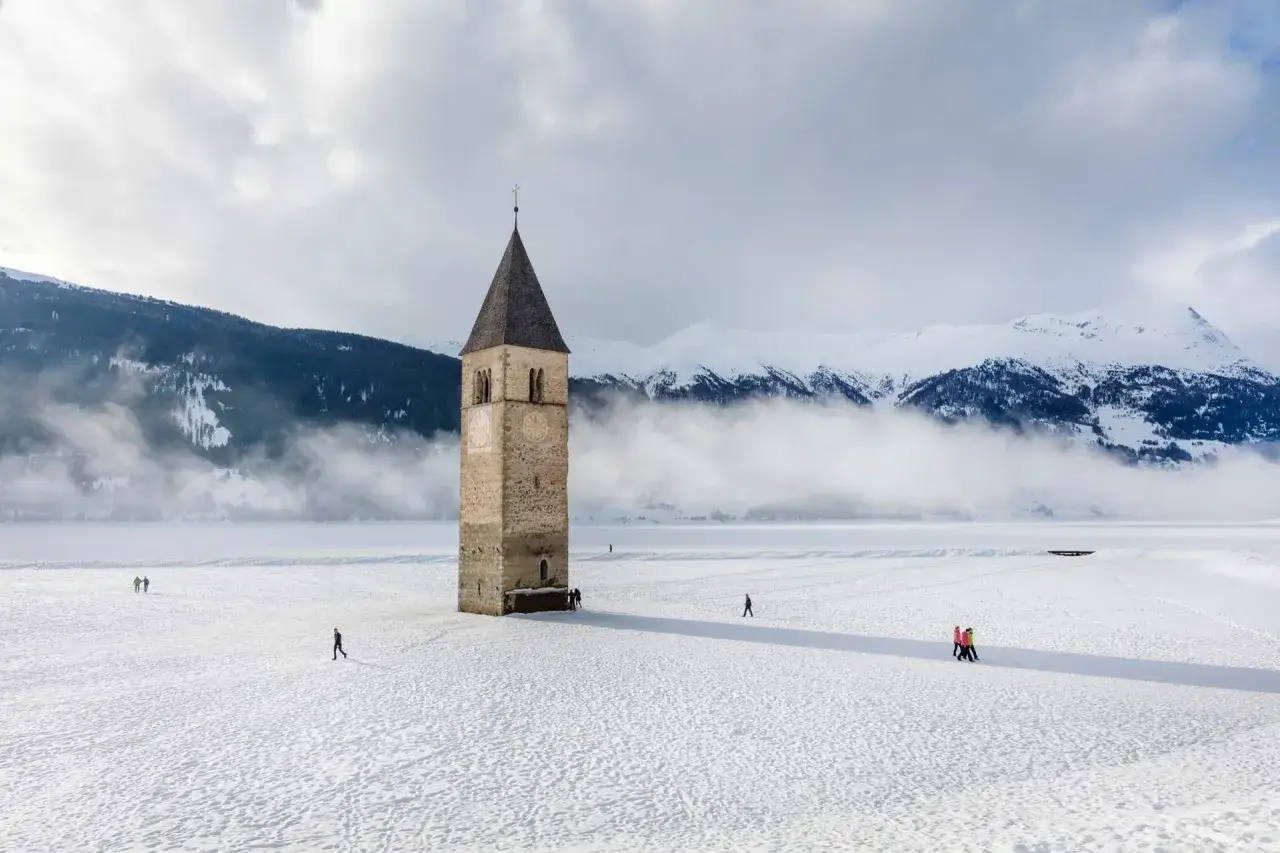 Il lago del campanile, con la sua torre che emerge dal ghiaccio, circondato da montagne innevate e nebbia.