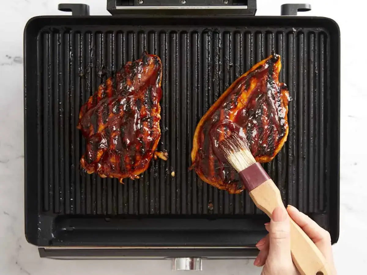 Dwa soczyste kawałki kurczaka w sosie barbecue są grillowane na elektrycznym grillu. Pędzelek właśnie nakłada kolejną porcję sosu. Dowiedz się, jak długo grillować kurczaka na grillu elektrycznym.