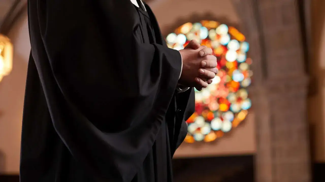 Ein Geistlicher in schwarzer Robe betet mit gefalteten Händen vor einem bunten Kirchenfenster. Er spricht das