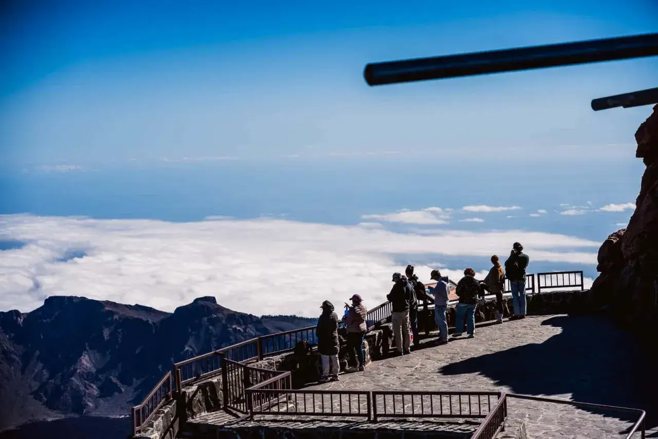 Vue depuis le pico mas alto de España, des touristes admirent la mer de nuages sous un ciel bleu éclatant.
