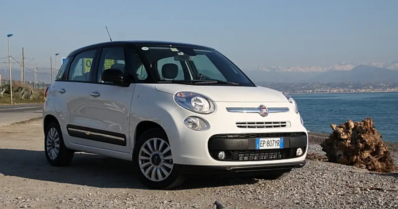 Biały Fiat 500 L z czarnym dachem stoi na kamienistej plaży nad morzem.