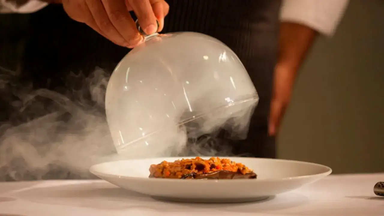 Plato ahumado servido en un restaurante con estrella Michelin. El humo escapa de una campana de cristal al ser levantada.