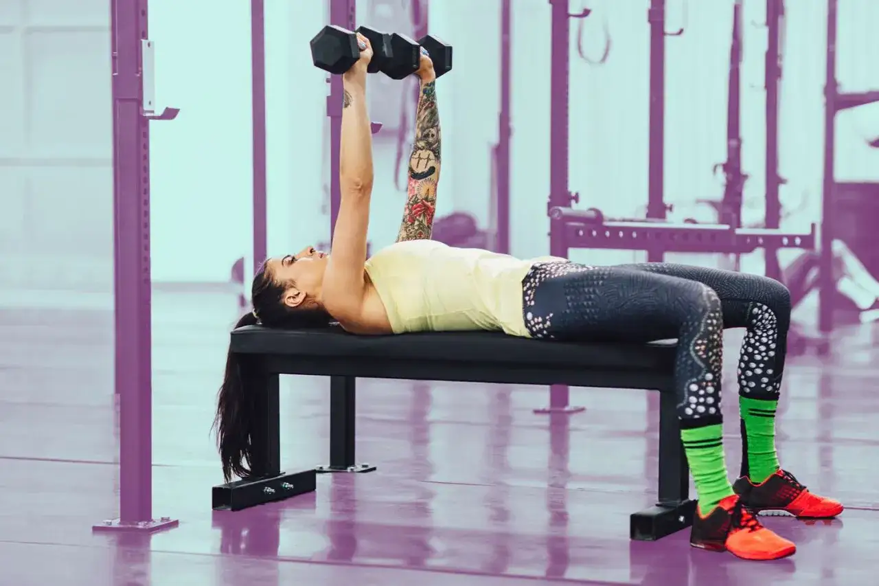 Kobieta wykonuje dumbbell press na ławce, z hantlami w dłoniach.
