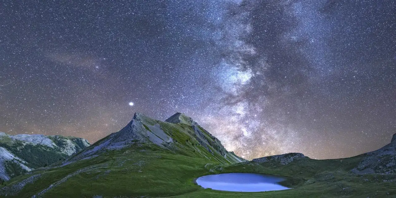 Le lac des Pises sous une voie lactée spectaculaire. Montagnes verdoyantes et ciel étoilé se reflètent dans l'eau paisible.
