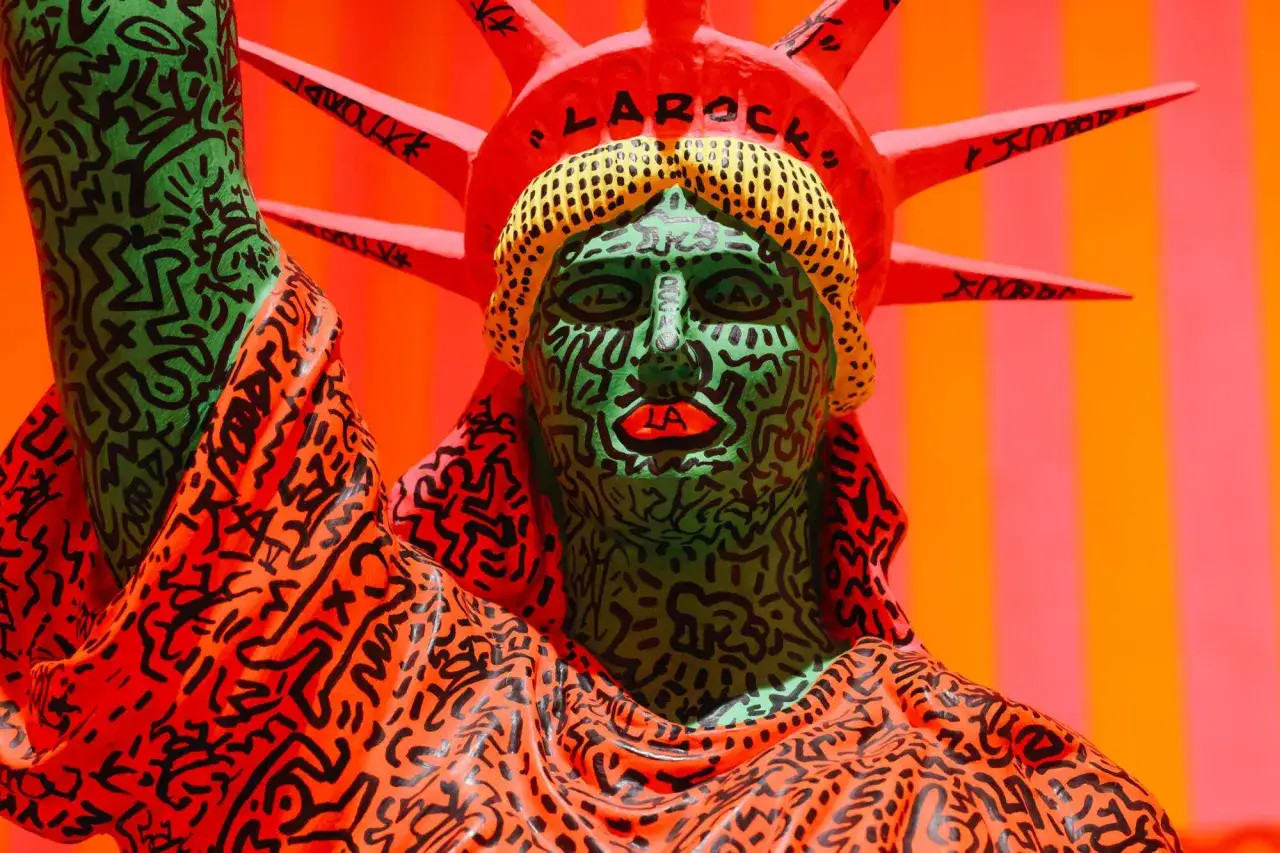 A vibrant Statue of Liberty sculpture, reimagined with Keith Haring's iconic queer art style, features bold black lines on a green face and orange robe.