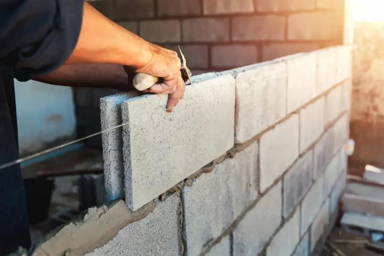 Un maçon pose une brique sur un mur en construction, indice bt 01. La lumière du soleil éclaire la scène.