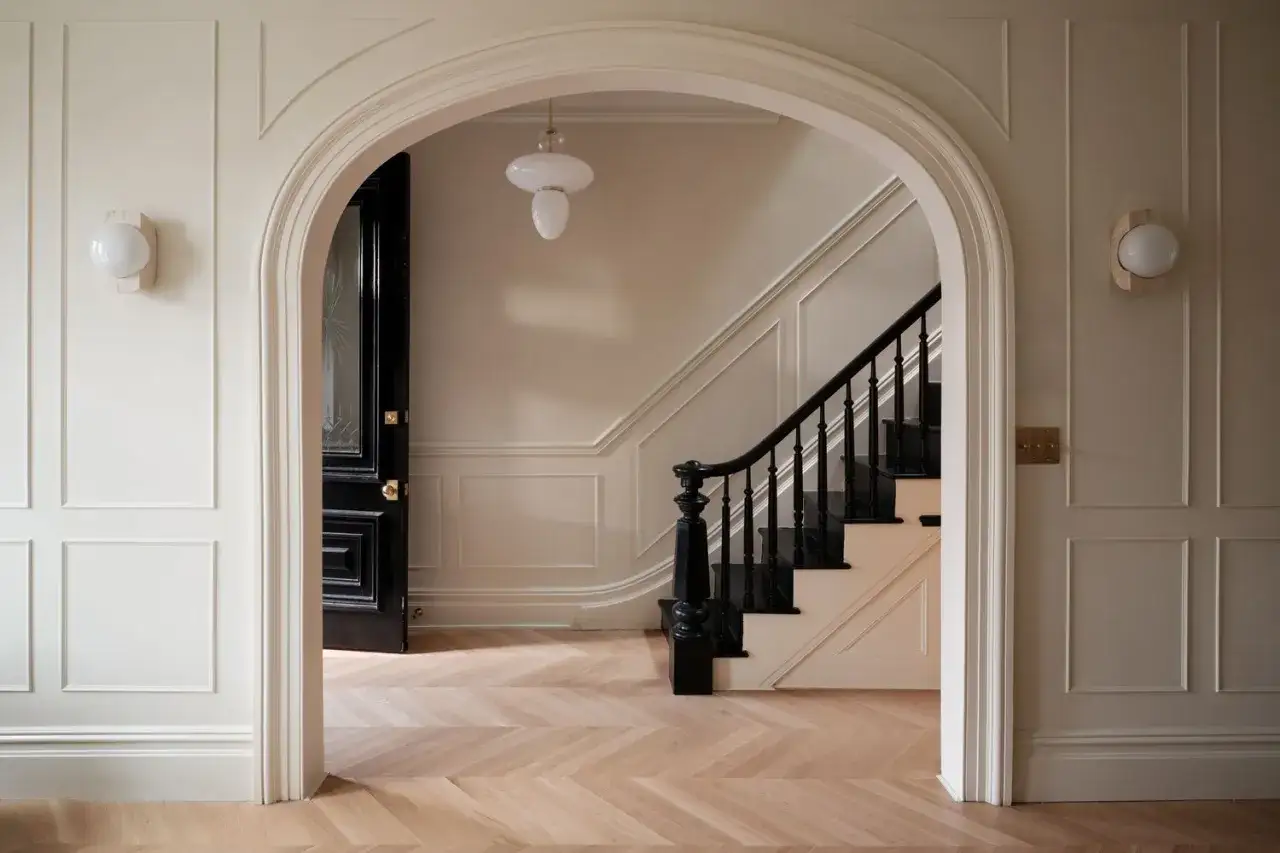 Entrada con puerta negra y escaleras, paredes con molduras y suelo de espiga. Colores suaves para pintar una casa, creando un ambiente acogedor.