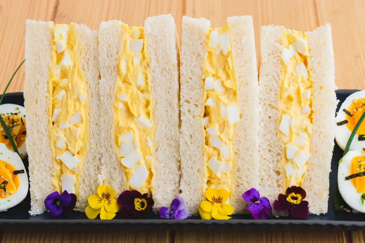 Quatre sandwichs tamago sando, garniture cr&eacute;meuse aux &oelig;ufs, pain blanc moelleux, d&eacute;cor&eacute;s de fleurs comestibles.