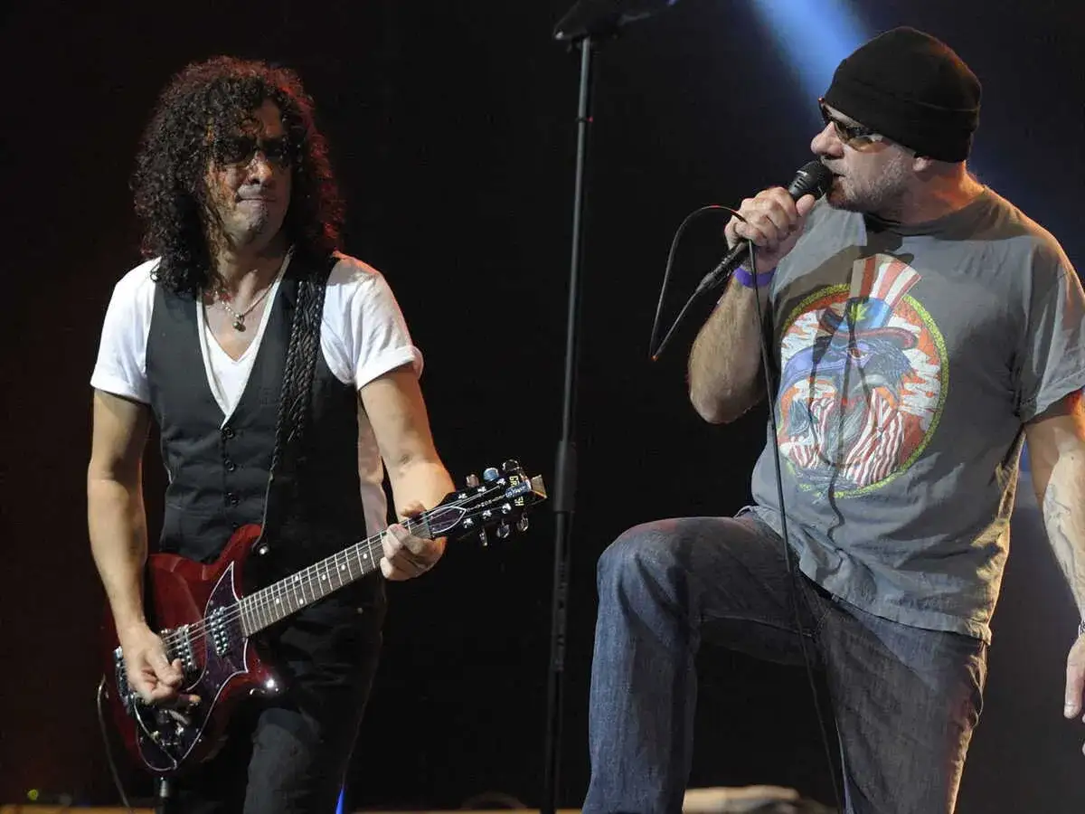 Deux membres de trust (groupe) sur scène : un guitariste aux cheveux bouclés et un chanteur au crâne rasé, casquette noire, lunettes de soleil et t-shirt graphique.