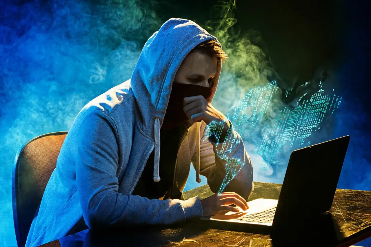 Mężczyzna w kapturze przy laptopie, otoczony cyfrowym dymem, szuka informacji, jak zablokować dowód osobisty.