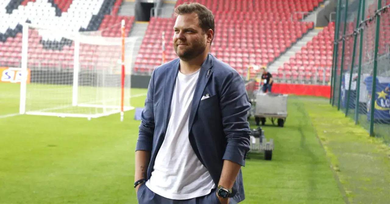 Jan Urban syn, w granatowym garniturze, stoi na tle stadionu.