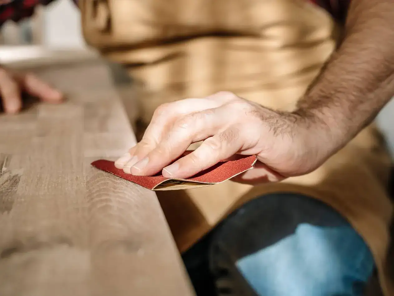 En hand slipar en tr&auml;yta med sandpapper. Det &auml;r en del av processen att slipa teak f&ouml;r att f&aring; fram dess naturliga sk&ouml;nhet.