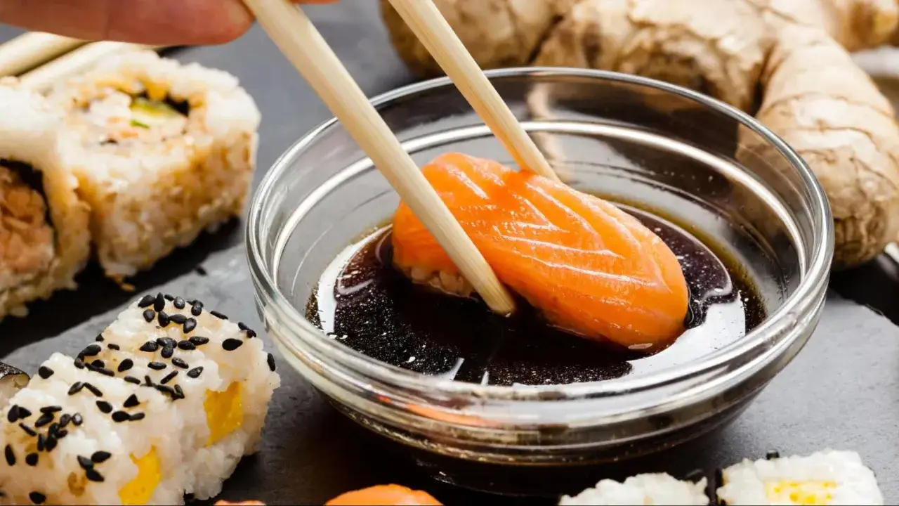 Kawałek łososia zanurzany w sosie do sushi. Obok sushi z sezamem i kawałek imbiru.