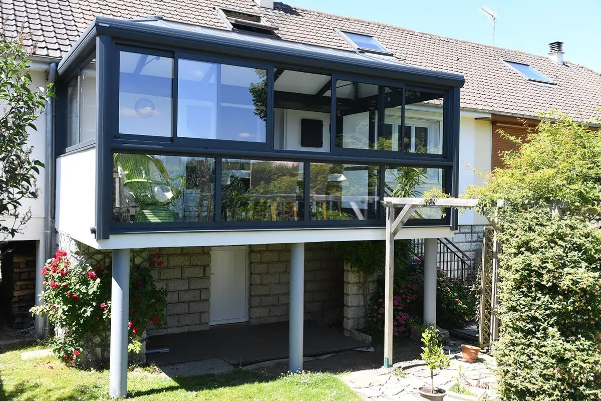 Veranda sur terrasse sur&eacute;lev&eacute;e, avec une chaise verte visible &agrave; travers les vitres. Le soleil brille sur le jardin verdoyant.