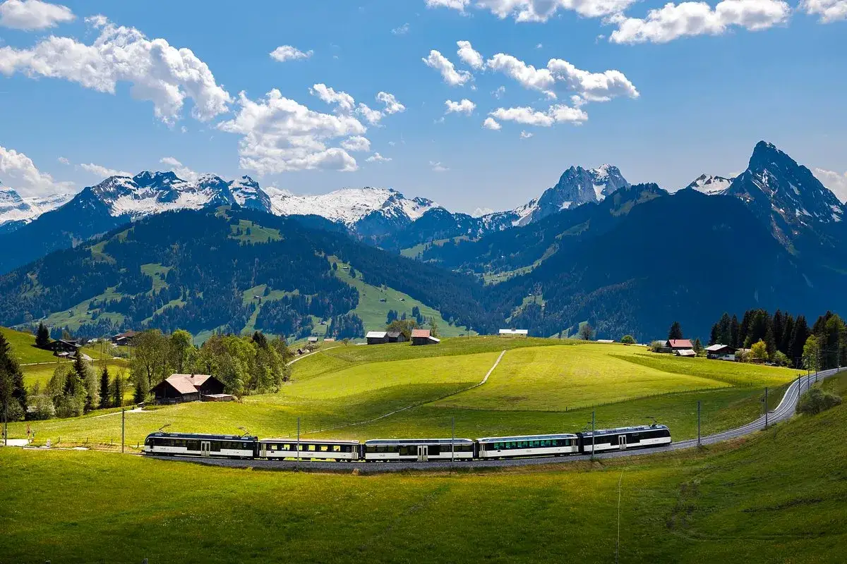 A train travels along the **Golden Pass Line** through a lush green valley, with snow-capped mountains in the background under a blue, cloudy sky.