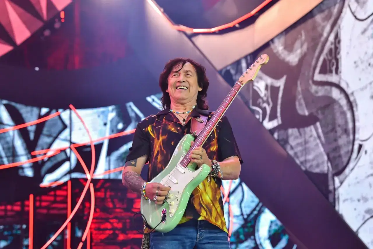Jan Borysewicz z Lady Pank gra na gitarze, rozpromieniony na tle sceny z efektami świetlnymi.
