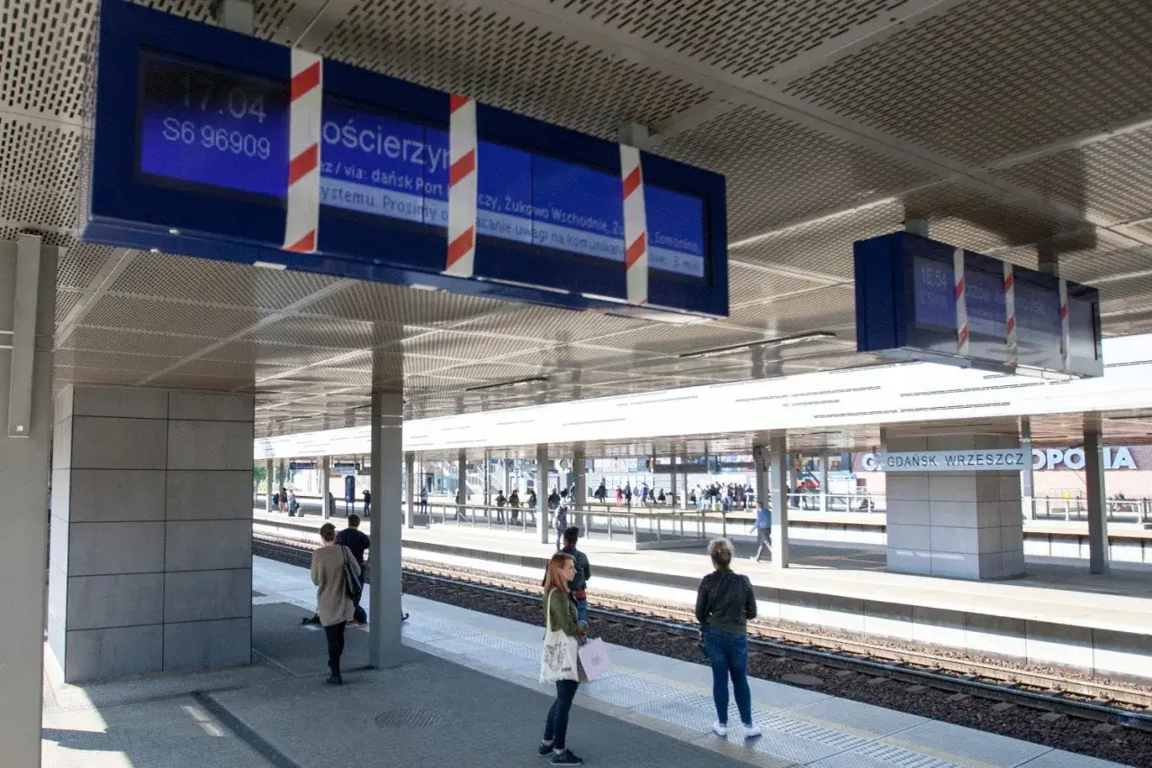 Na tablicach informacyjnych na dworcu kolejowym widać, jak sprawdzić z którego peronu odjeżdża pociąg. Ludzie czekają na peronach.