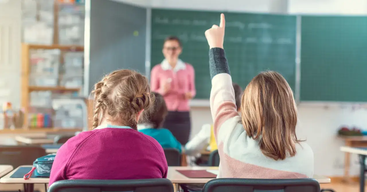 Uczennica podnosi rękę, by odpowiedzieć na pytanie. W tle widać nauczycielkę przy tablicy, która z uśmiechem tłumaczy lekcję.