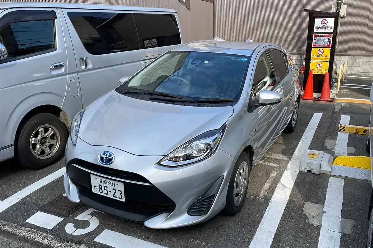 Srebrny kei car zaparkowany obok vana. Na tablicy rejestracyjnej widnieje napis