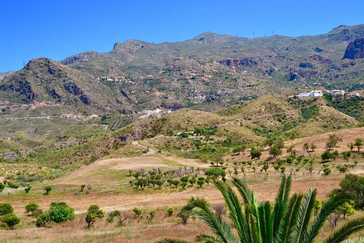 Un cortijo grande se alza en una colina, rodeado de un paisaje montañoso árido y vegetación dispersa bajo un cielo azul intenso.