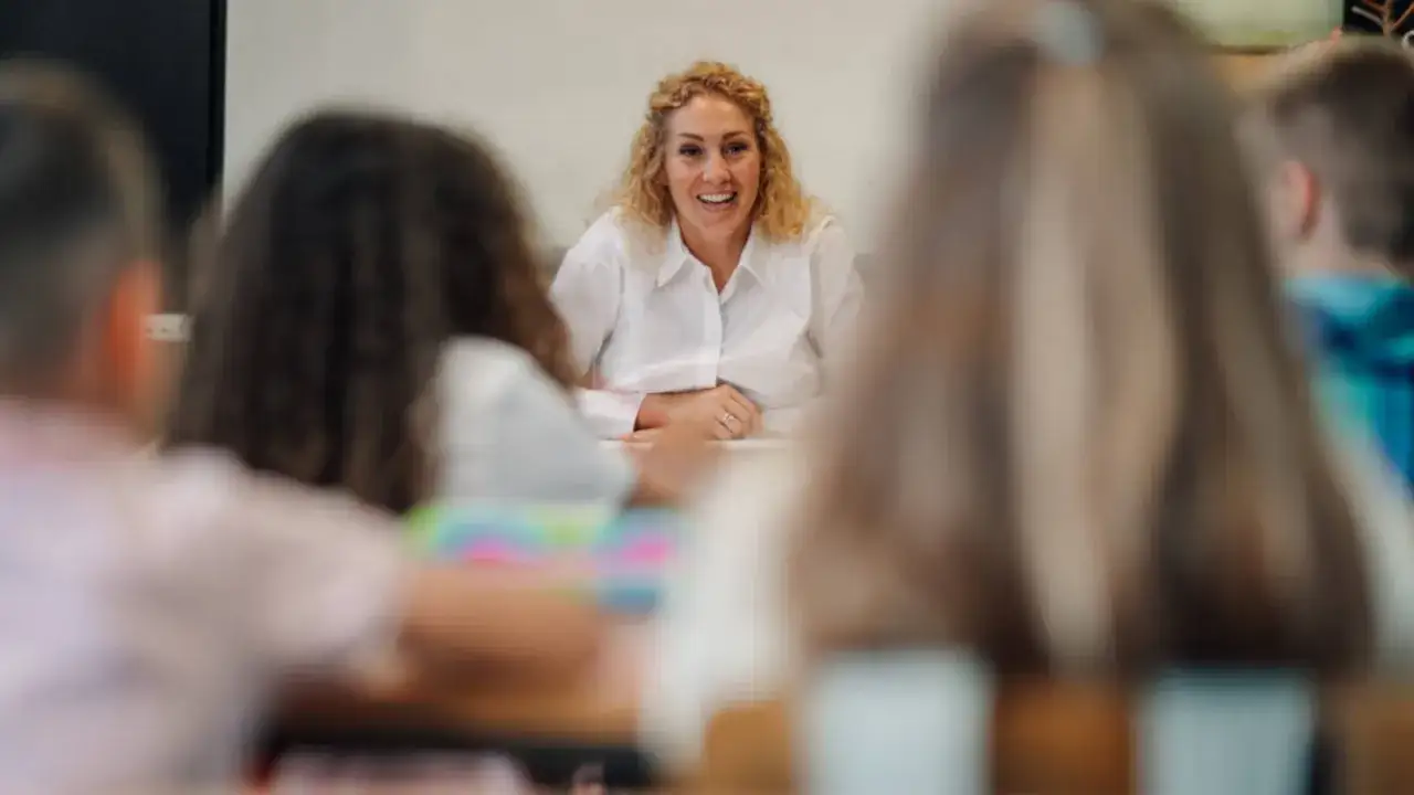Uśmiechnięta nauczycielka prowadzi zajęcia, które mogą być częścią przygotowania pedagogicznego dla psychologów. Dzieci siedzą w ławkach.