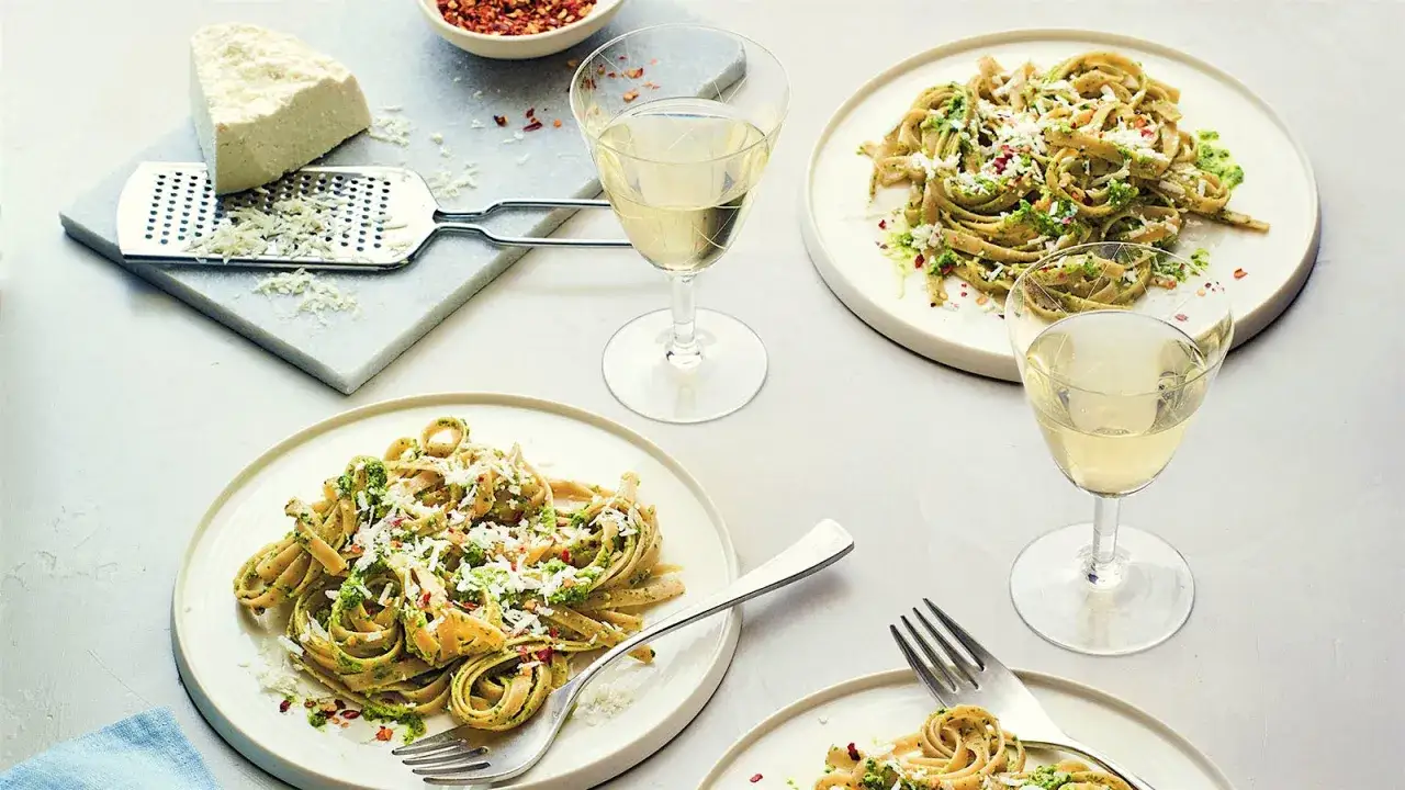 Två tallrikar med pasta pesto, toppad med riven ost och chiliflakes. Ett glas vitt vin till varje portion. Perfekt för en mysig middag.