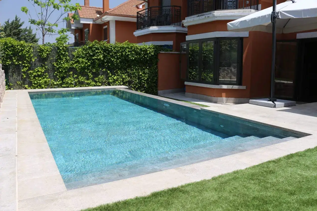 Una moderna piscina desbordante con agua turquesa, rodeada de césped y una casa de color terracota.