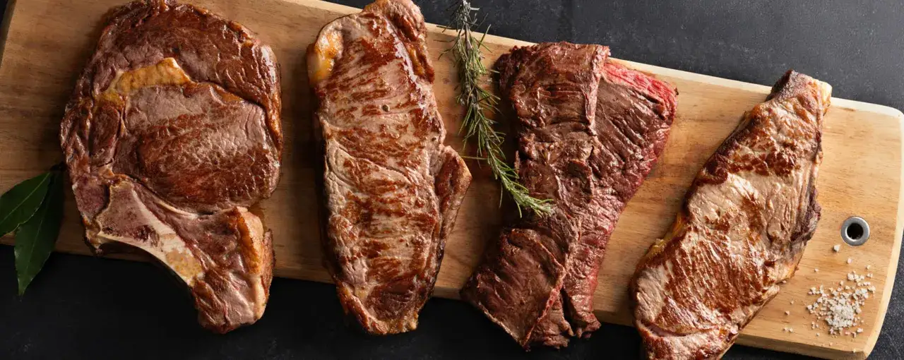 Quatre pi&egrave;ces de viande grill&eacute;e, dont un faux filet, dispos&eacute;es sur une planche en bois avec des herbes et du sel.