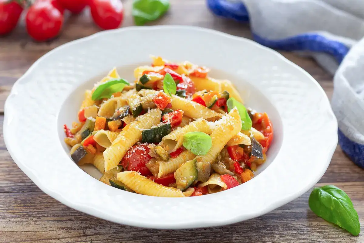 Piatto di pasta con verdure miste, pomodorini, zucchine e basilico fresco, guarnito con parmigiano grattugiato.