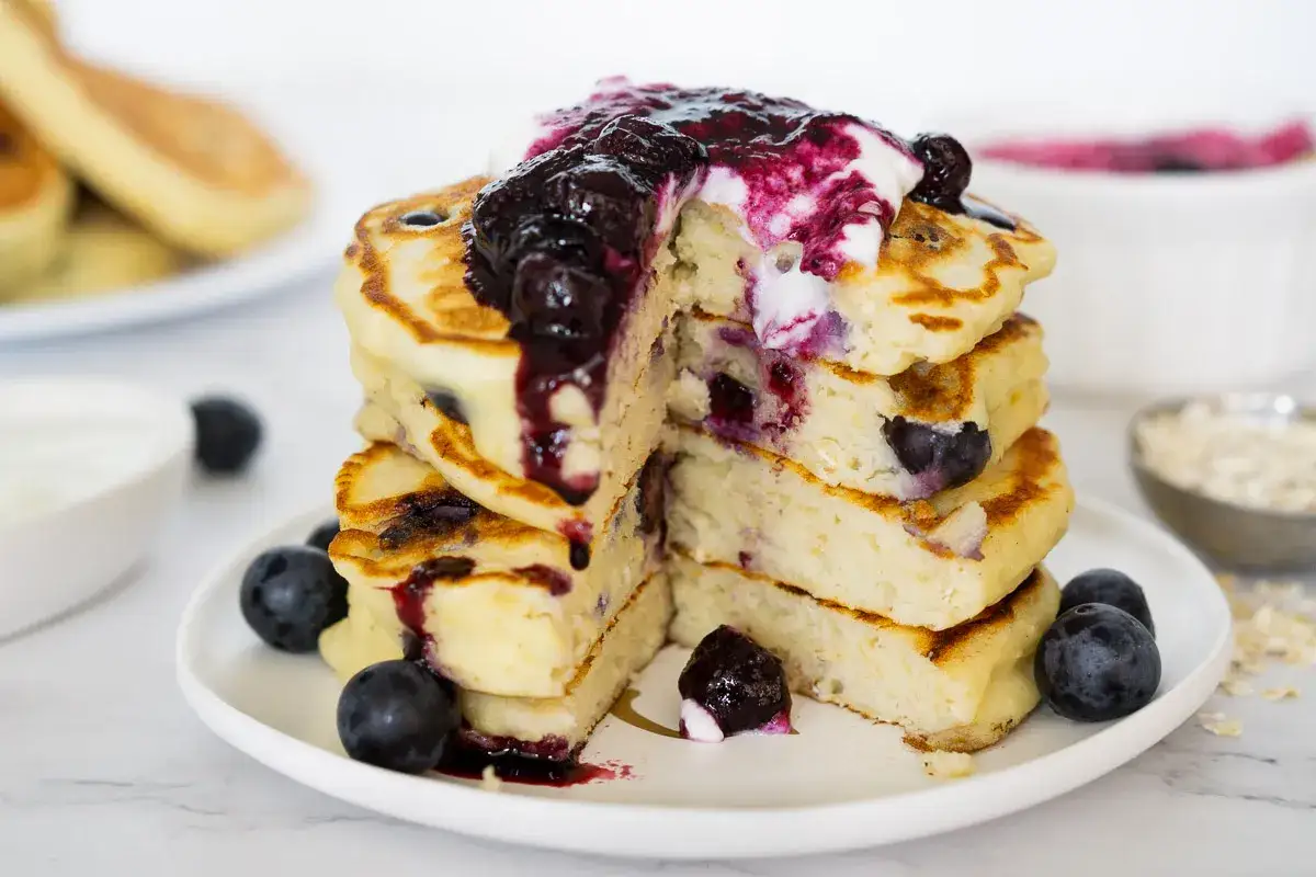 En hög med fluffiga, nyttiga pannkakor med blåbär och en klick grädde toppad med blåbärssås. Perfekt för en hälsosam frukost.