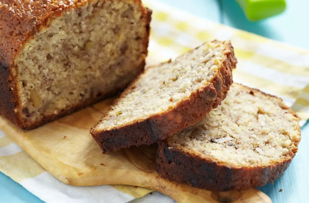 Rebanadas de delicioso banana bread casero, con nueces visibles, sobre una tabla de madera y un paño a cuadros.