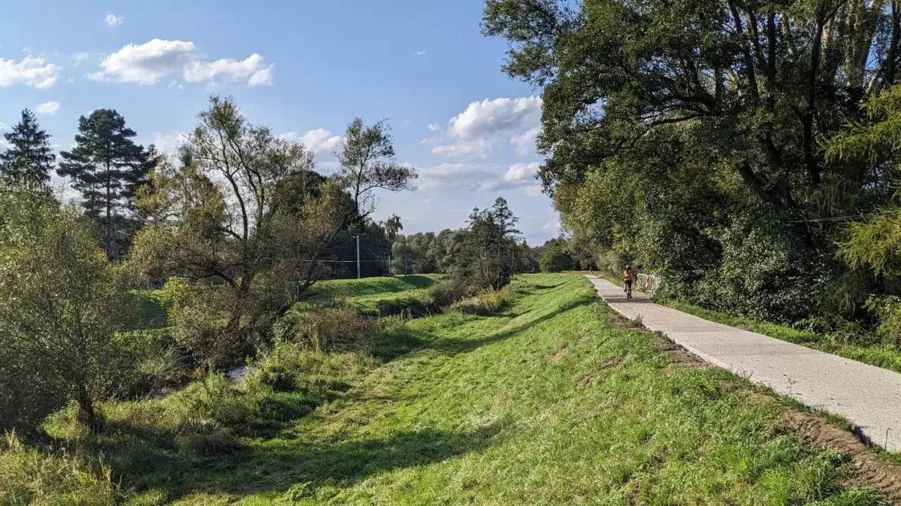 Osoba na rowerze jedzie ścieżką wzdłuż zielonej doliny. W oddali widać las, a na niebie chmury. To idealne miejsce na przejażdżkę velo rudawa.