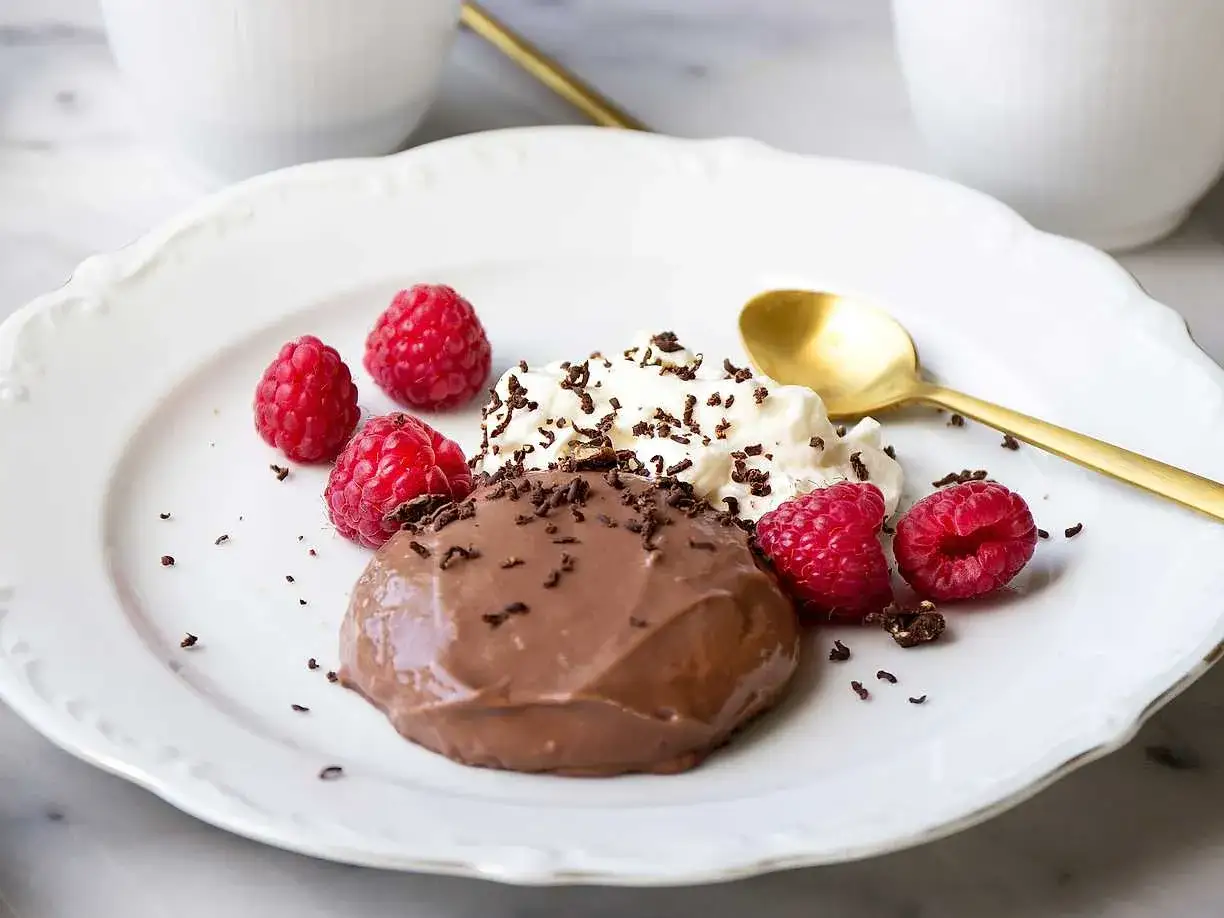 En härlig hemmagjord chokladpudding med vispgrädde, hallon och chokladströssel på en vit tallrik. En gyllene sked ligger bredvid.