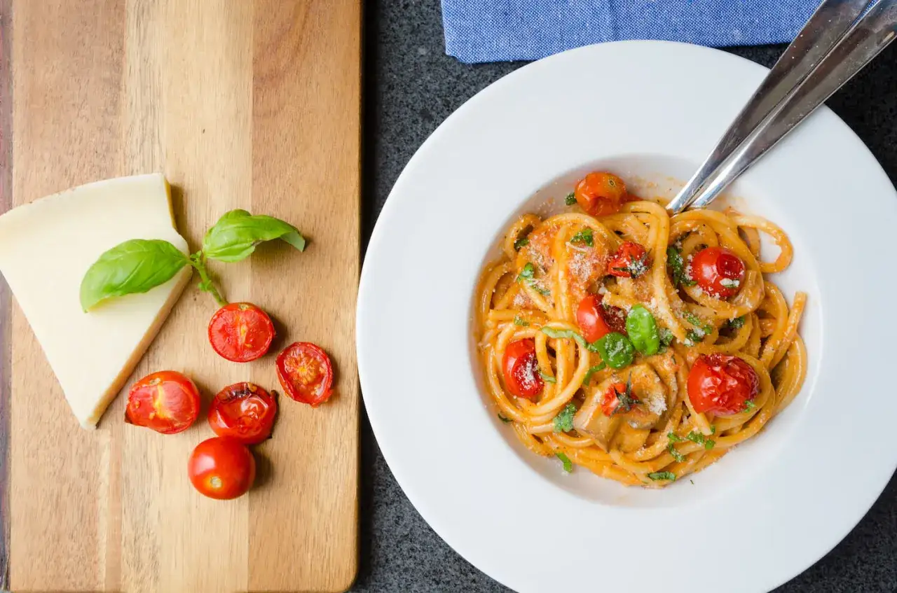 En tallrik med god pasta med tomater och basilika. Bredvid ligger en bit ost och några körsbärstomater på en träskärbräda.