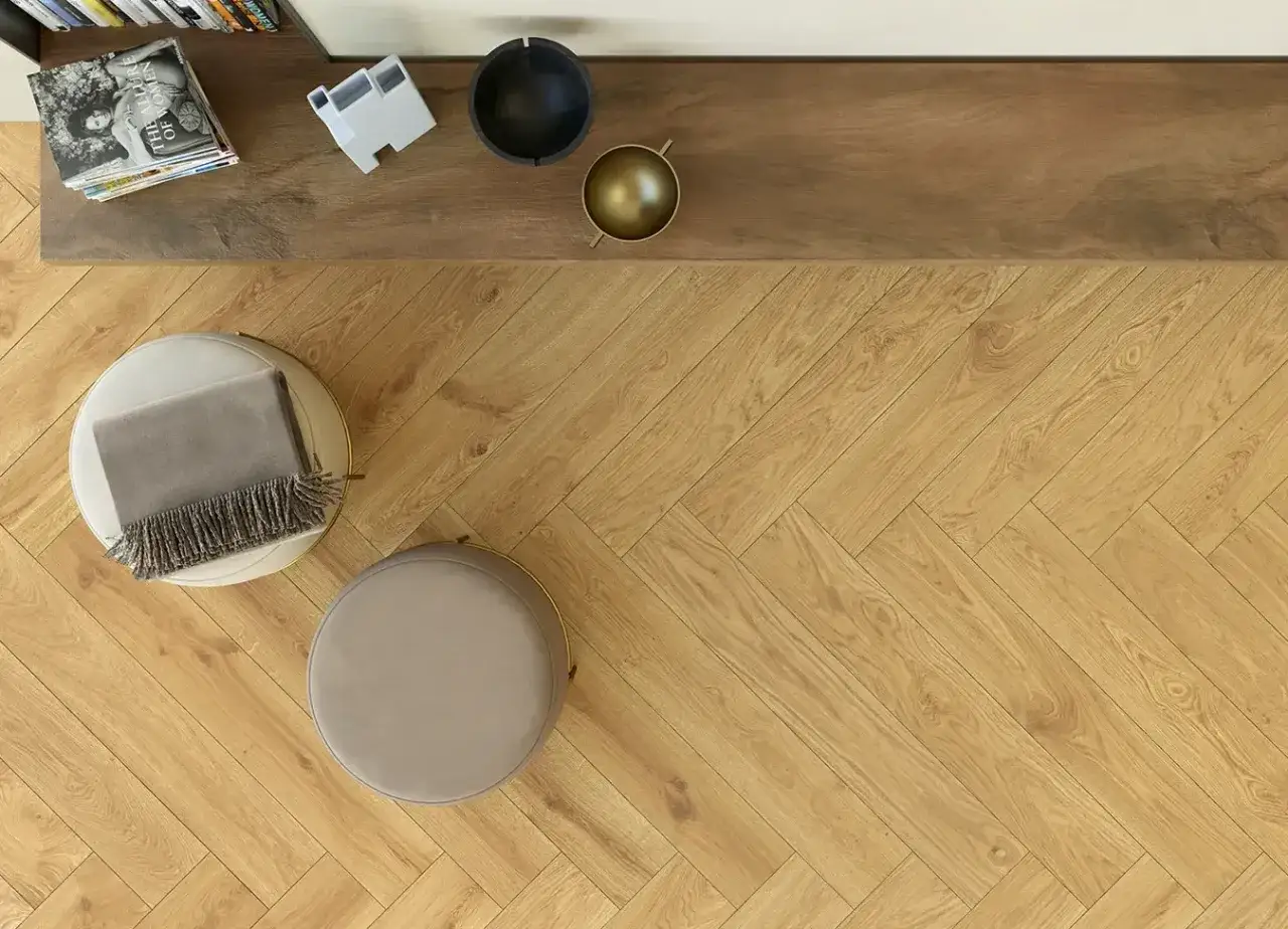 Schema posa piastrelle a spina di pesce su un pavimento in legno chiaro. Due pouf e una mensola con oggetti decorativi completano la scena.
